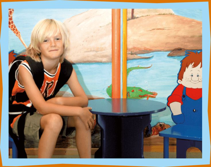 Blonde boy with black backpack, orange tank top and green shorts sitting on a chair.