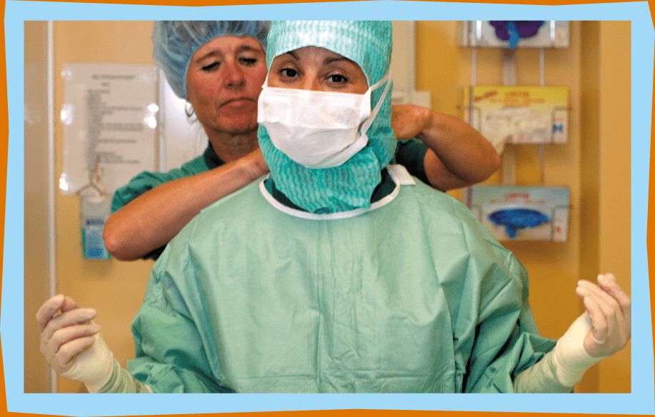 Surgeon in surgical scrubs, latex gloves, surgical cap and mask stands in front of nurse who helps her get dressed.