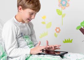 A boy dressed in white operating clothes is sitting in a hospital bed playing with his Ipad.