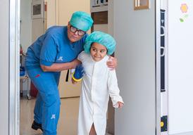 A nurse in blue scrubs and a little girl in white operating clothes are entering the door to an operating room. Both the nurse and the girl look very curious.