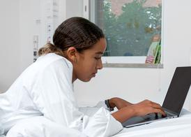 A teenage girl lying on her stomach in her hospital bed looking at her computer.