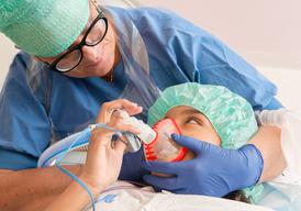 A girl is lying on an operating table with a face mask over her mouth. She is looking at a nurse who is sitting behind the table.