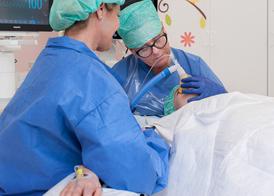 A patient is lying on an operating table breathing in an oxygen mask. The patient’s mother is sitting beside the table.