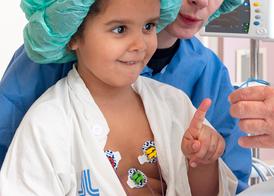 A girl in a green scrub cap sitting in her mother’s lap in an operating room. She is reaching out a finger to the oxygen meter and looks interested and curious.