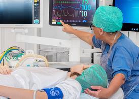 A nurse sitting behind an operating table where a patient is lying. The patient and the nurse are looking at numbers at the monitors behind the nurse.