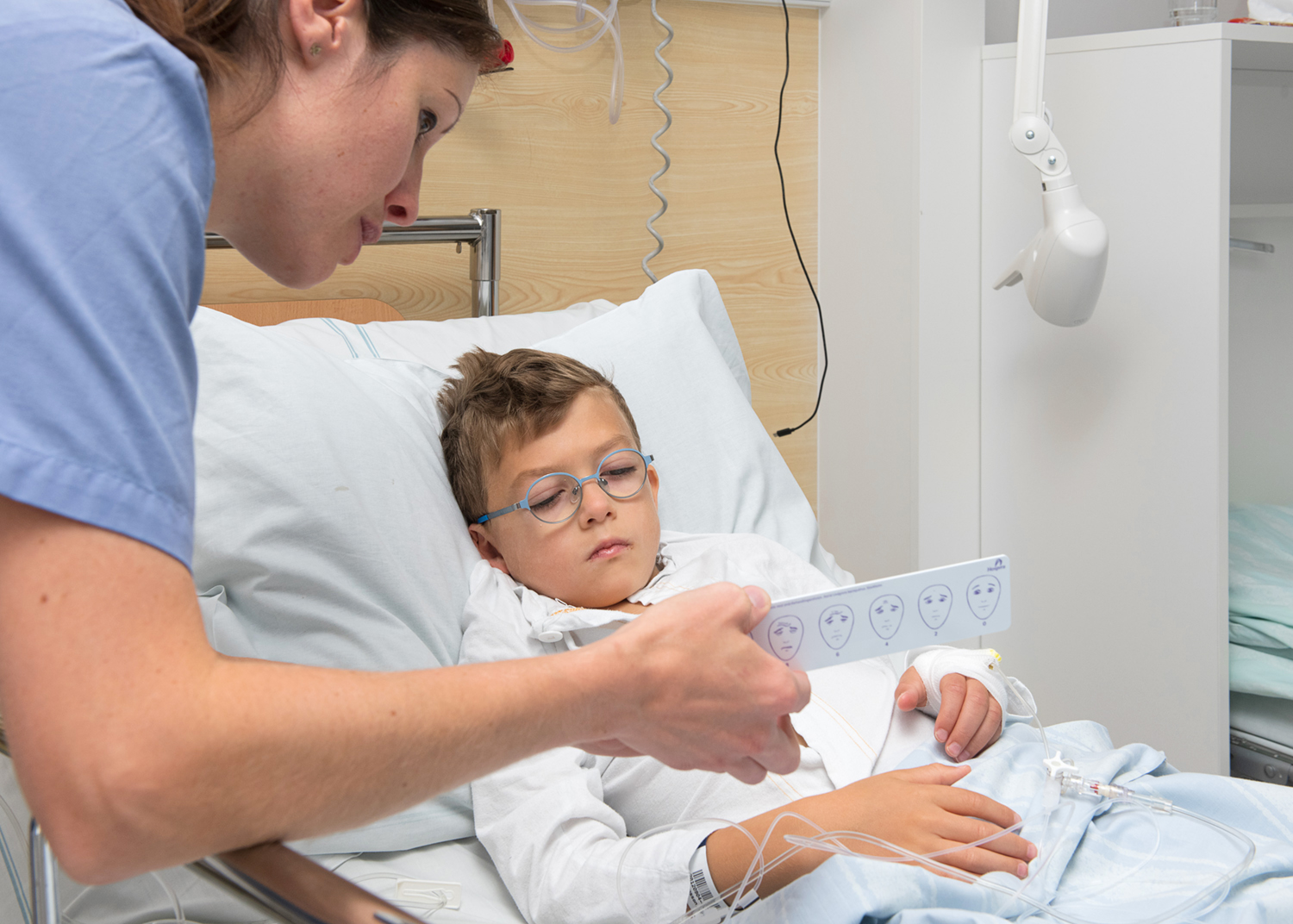 Nurse holding a ruler with faces on in front of a boy lying in bed.